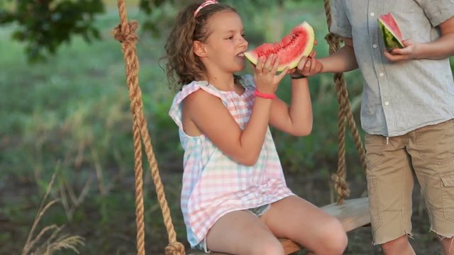 Happy girl and boy eating a watermelon sitting on a swing, summer photo. Children with a watermelon