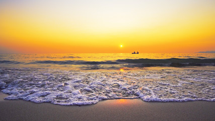 Small boat of refugees sail on sea horizon against sunset seen from beach splashed by waves