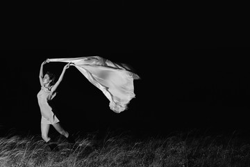 Amazing girl dancing in dress with big white shawl in darkness in grayscale. Beautiful female dancer with good mood in night. Unimaginable mystic dance on nature in dark in monochrome.