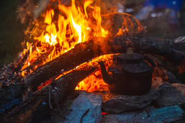 Boiling of tea in kettle on bonfire with large firewood. Tea drinking in open air. Active outdoor recreation. Camping in dusk. Romantic warm atmosphere in twilight on nature. Active rest.