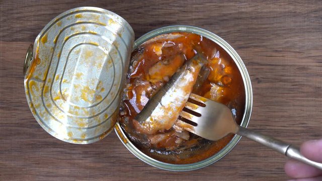 Using fork takes a sprats fish, sardines in tomato sauce from a tin can. Top view, close up, macro