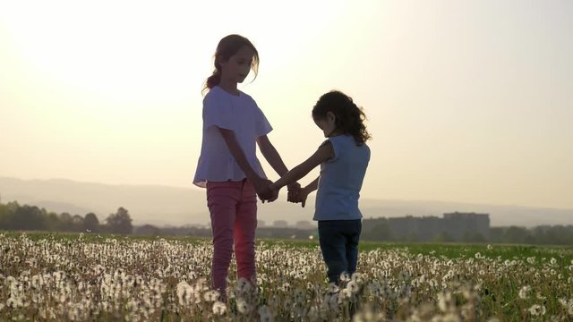 Two sisters holding hands circling on the glade of dandelions in the city park outdoors. Freedom and carefree. Happy childhood.