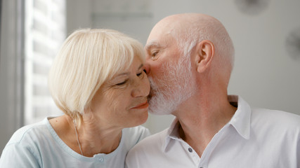 Portrait of happy senior couple at home. Senior man expresses his emotions and kisses his wife. Very emotional moment. Happy family enjoying time together.