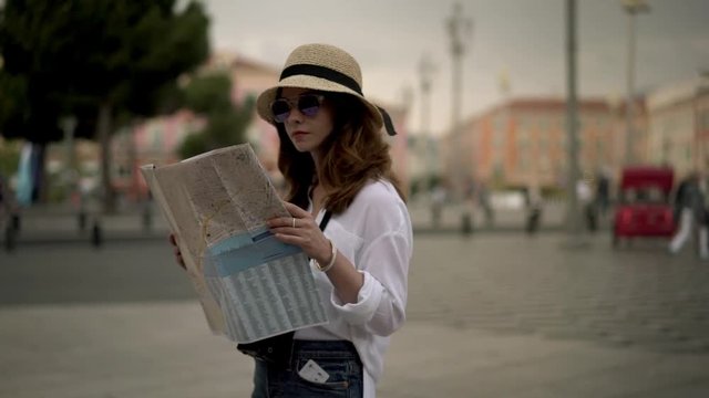 Attractive young woman tourist in purple sunglasses and straw hat looking around while holding large map standing in French city of Nice on sunny spring day. Handheld slow motion medium shot