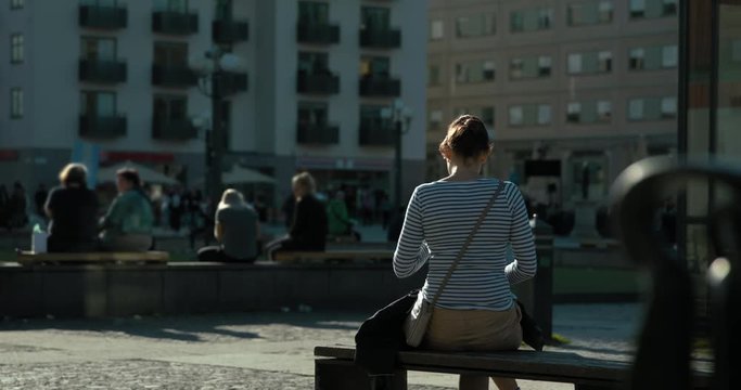 Slow motion, woman using phone and relaxing on a bench in a Stockholm square. Blurred people in the background.
