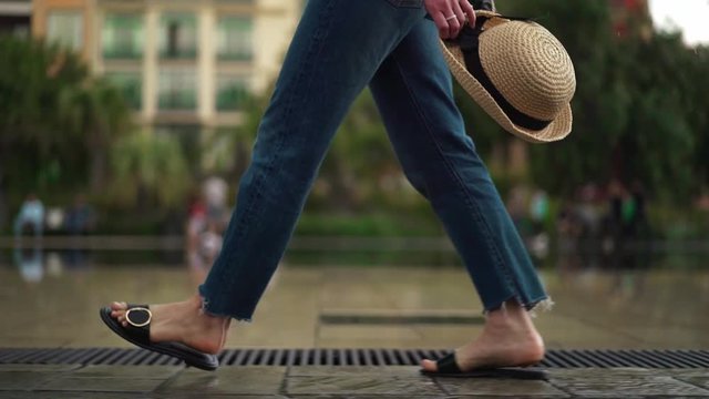 Side view of unrecognizable woman legs in jeans and sandals walking along a fountain in Nice, France and holding her straw hat. Tracking real time medium shot