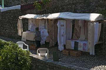 Bosnia: un'anziana donna bosniaca di spalle che vende tessuti e pizzi finemente ricamati, creati secondo l'antica tradizione balcanica nei vicoli di Mostar