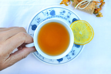  Senior woman holding cup of hot tea with lemon slice.Rustic porcelain cup a herb wrapped up as a gift on a white tablecloth with lace.Top view. Healthy concept. 