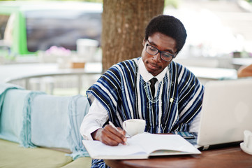 African man in traditional clothes and glasses sitting at outdoor caffe, drink coffee behind laptop and writing something on his notebook.