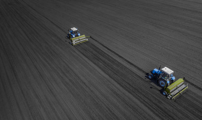 Obraz premium Aerial view of two blue tractors plows the earth in field on a summer day against a black earth background. Agriculture. Two tractors travel one after another along the black field