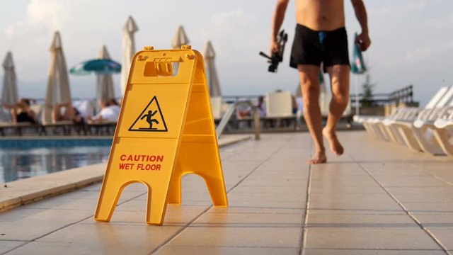 Barefoot man walks toward camera with caution wet floor sign near the swimming pool