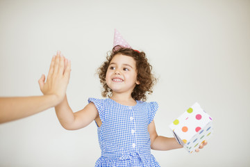 Pretty little girl with dark curly hair in blue dress and birthday cap happily looking aside while giving high five with gift box in hand over white background