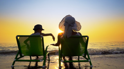 Fototapeta na wymiar Happy family enjoy luxury sunset on the beach during summer vacations. Mother and son are sitting on a beach deck chair, against sunset, cinematic steadicam shot