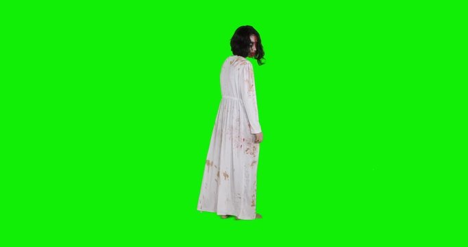 Creepy zombie woman with wounded face and white gown standing in the studio, shot in 4k resolution with green screen background