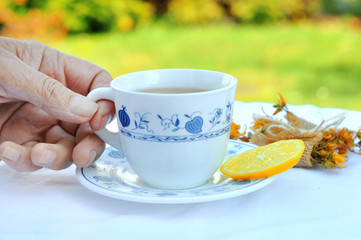 Senior woman holding cup of hot tea with lemon slice in nature.Rustic porcelain cup a herb wrapped up as a gift on a white tablecloth with lace. Healthy concept. 