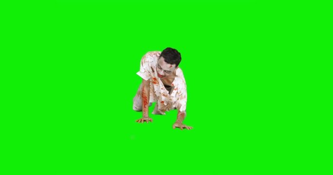 Horrible scary zombie man with bloody mouth and torn clothes, crawling in the studio against green screen background. Shot in 4k resolution