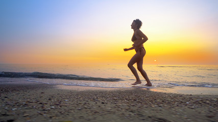 Fototapeta na wymiar Barefoot sport woman in bikini jogging, running on an empty beach at sunset