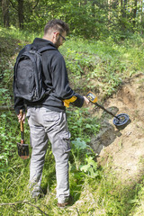 Mężczyzna z plecakiem na plecach stoi w lesie z wykrywaczem metali w ręce i przeszukuje teren w poszukiwaniu skarbów.