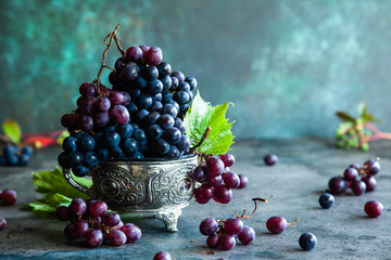 Red and black grapes with grape leaves in a metal bowl on a rustic background.