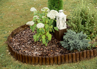 Round flower bed covered by pine bark mulch and surrounded by brown weatherproof wood roll boarder palisade, inside are different plants, outdoors in summer. - obrazy, fototapety, plakaty