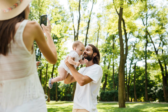 Young woman in a hat takes a photo of her husband holding in the arms their little charming daughter on a sunny day in the park.