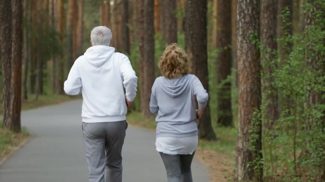 Rear view of unrecognizable elderly woman and man jogging along pavement road in park