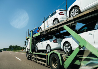 View of lgeneric white long truck trailer transporting new cars and riding on highway in sunlight under blue sky