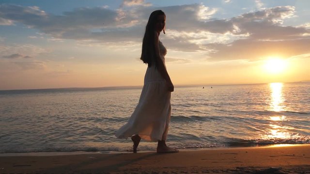 young woman in sundress walking on the beach at sunset