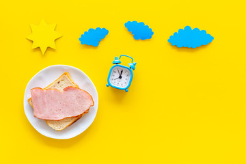 Parts of day. Morning. Time for breakfast. Sandwich near alarm clock, sun and clouds cutout on yellow background top view copy space