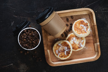 Wooden serving tray with curd fritters and takeaway coffee for breakfast, top view on a dark brown stone background