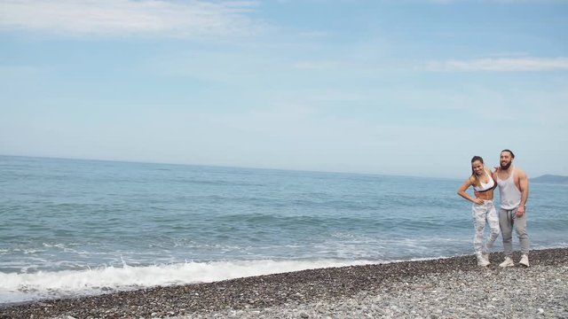 Happy sportive friends, young bearded man with haik knot and attractive fit woman relaxing on the seashore, having rest after intense crossfit workout. Vacation SportLife, Honeymoon Getaway.