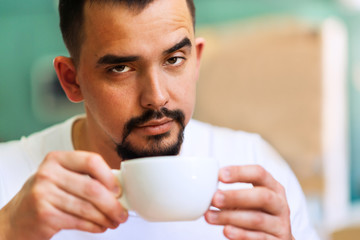 Handsome man with raised eyebrow and ironical face looking at camera over the white hot mug of morning coffee. Morning coffee ritual. Room for text