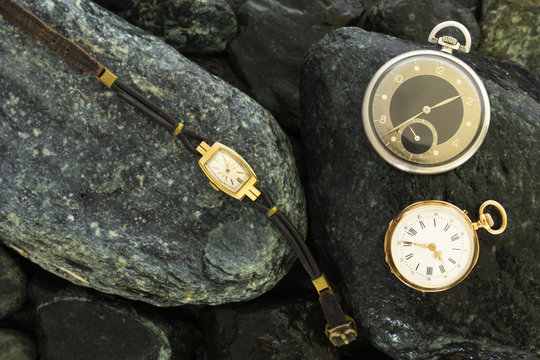 Set of watches on wet green stones with a classic gold pocket watch a black and silver pocket watch and a wristwatch with worn leather straps