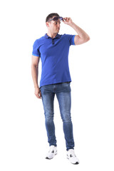 Fototapeta premium Confident young macho adult man in blue polo shirt holding and looking away under sunglasses. Full body isolated on white background.