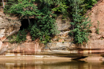 Fototapeta na wymiar sunny summer day at the river and the red rocks of the cliff grow some trees, rock reflection in the river
