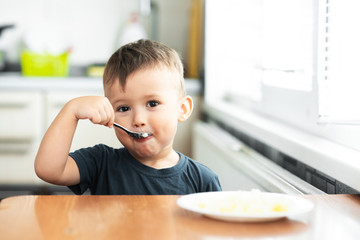 The little boy in the kitchen eagerly eating rice with a spoon independently