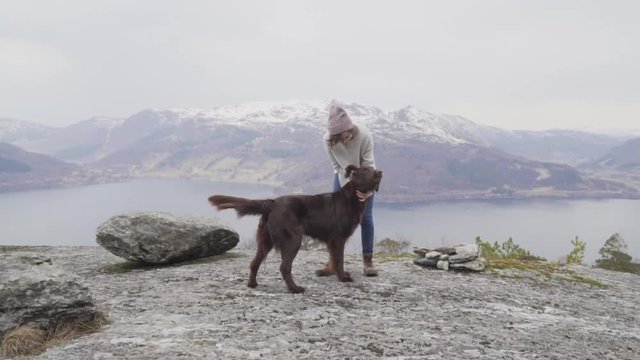 Woman with glasses, red cap, grey sweater, blue jeans and brown boots playing and petting a chocolate flat coated retriever on the top of a mountain with a view to the fjord and snowy mountains_slomo