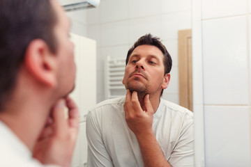 Morning hygiene, man in the bathroom looking in mirror and and checks his beard.