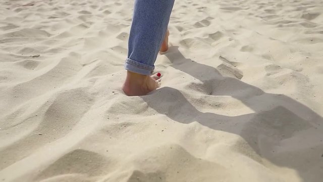 Rear view of woman feet barefoot in jeans walking on sandy beach. rest by the sea on the Golden sand. Low camera angle and slowmotion.