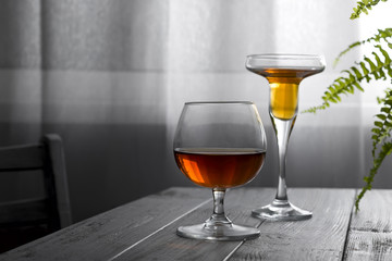 two glasses of wine placed on a wooden table on the background of a window with a white curtain. Natural light from a window in a dark room with a flower in the pot. Copy space