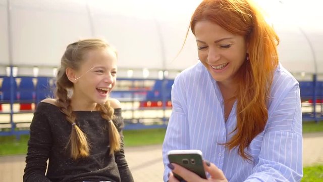 young mom and her daughter are watching a hilarious smartphone video and smiling while sitting in the Park for a walk