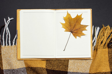 Opened notebook, yellow autumn maple leaf, orange brown checkered plaid on black background top view copy space. Autumn flat lay background. Autumn, fall, halloween concept.