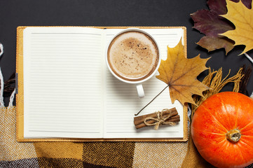 Opened notebook, cup of pumpkin cocoa or coffee, cinnamon, yellow autumn maple leaf, orange brown checkered plaid on black background top view copy space. Autumn flat lay background