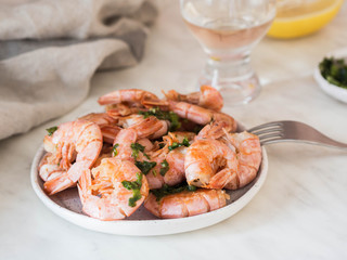 White plate with prawns fried with herbs and garlic and glass and white wine on a white table