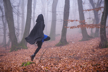 Girl in black hood running away from danger deep in dark forest. Thick fog all around. Scary autumn scene
