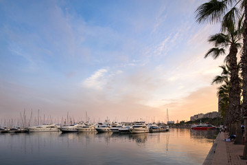 Fototapeta premium Beautiful port of Alicante, Spain at Mediterranean sea. Luxury yachts, ships, ferries and fishing boats sailing and standing in rows in harbor. Rich people traveling around the world. Sunset evening
