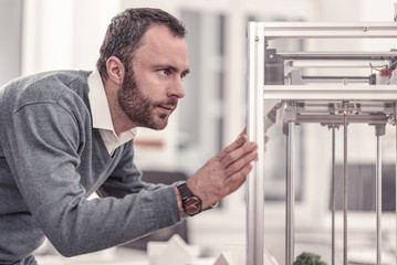 Fascination. Clever bearded man watching at 3D printer waiting when his prototype being ready