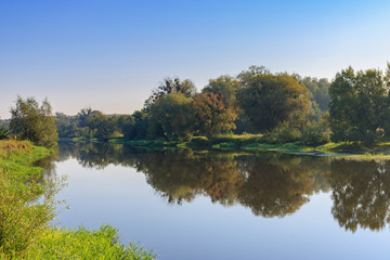 Fototapeta na wymiar River landscape in sunny summer morning on a background of green trees on the shore