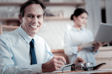 Happy man. Positive successful businessman feeling glad and smiling while sitting at the table and...