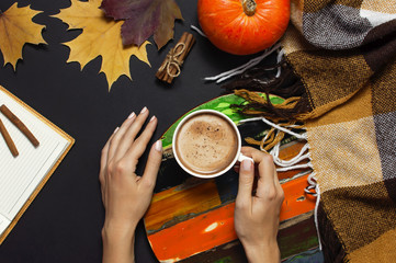 Cup of pumpkin cocoa or coffee in female hands, opened notebook, pumpkin, cinnamon, yellow autumn maple leaf, checkered plaid on black background top view copy space. Autumn flat lay background